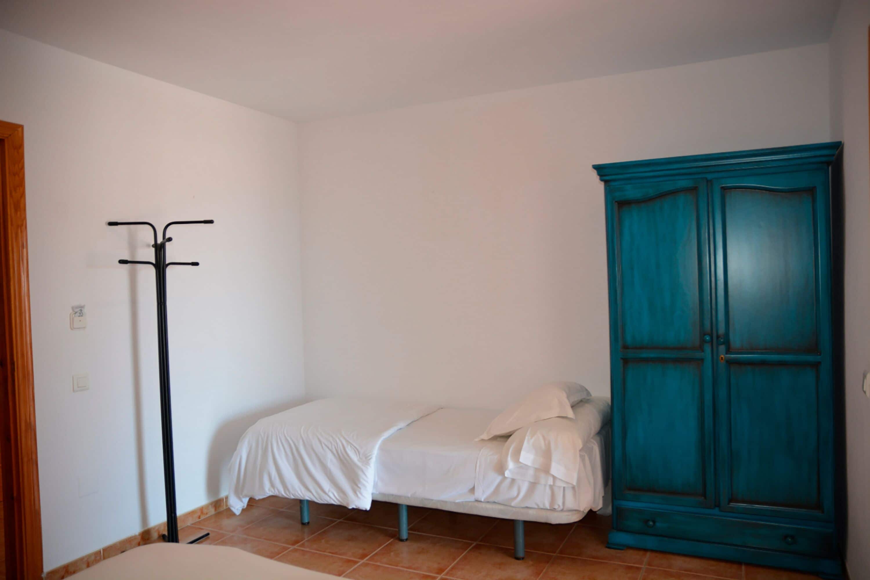 Ein kleines Zimmer mit einem Einzelbett, einem türkisfarbenen Kleiderschrank und einem Kleiderhaken.