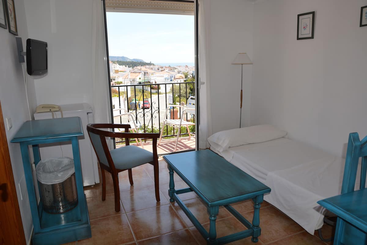 A room with a bed, a table, a chair, a trash bin, and a view of the outside.