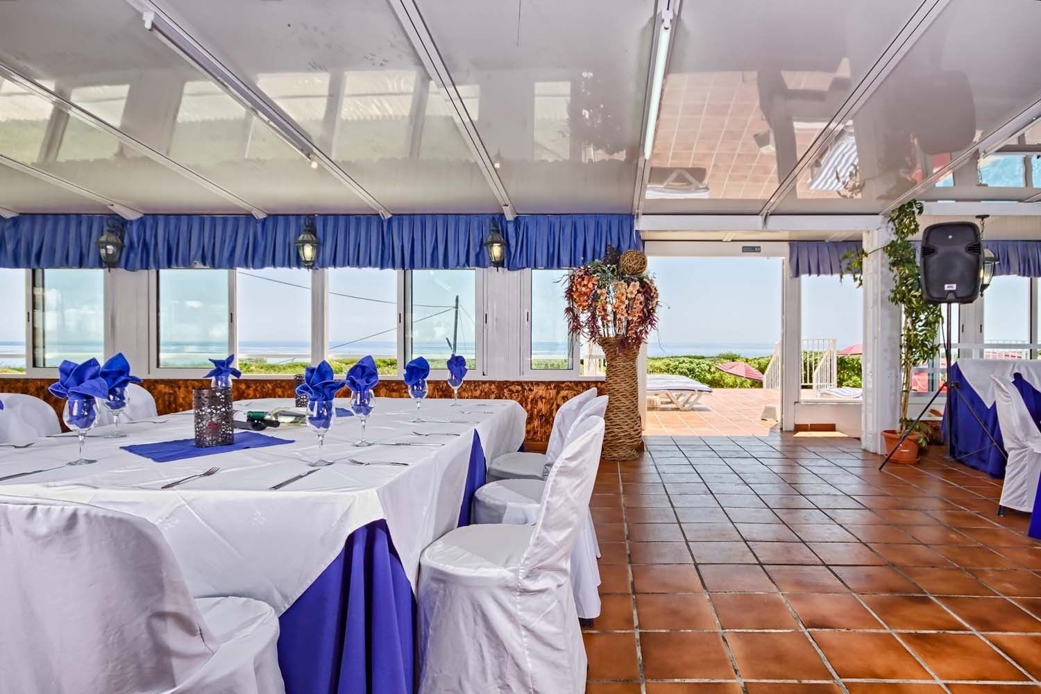 Una sala da banchetto con tavoli apparecchiati per un evento formale, ampie finestre vista mare e un palco con un oratore.