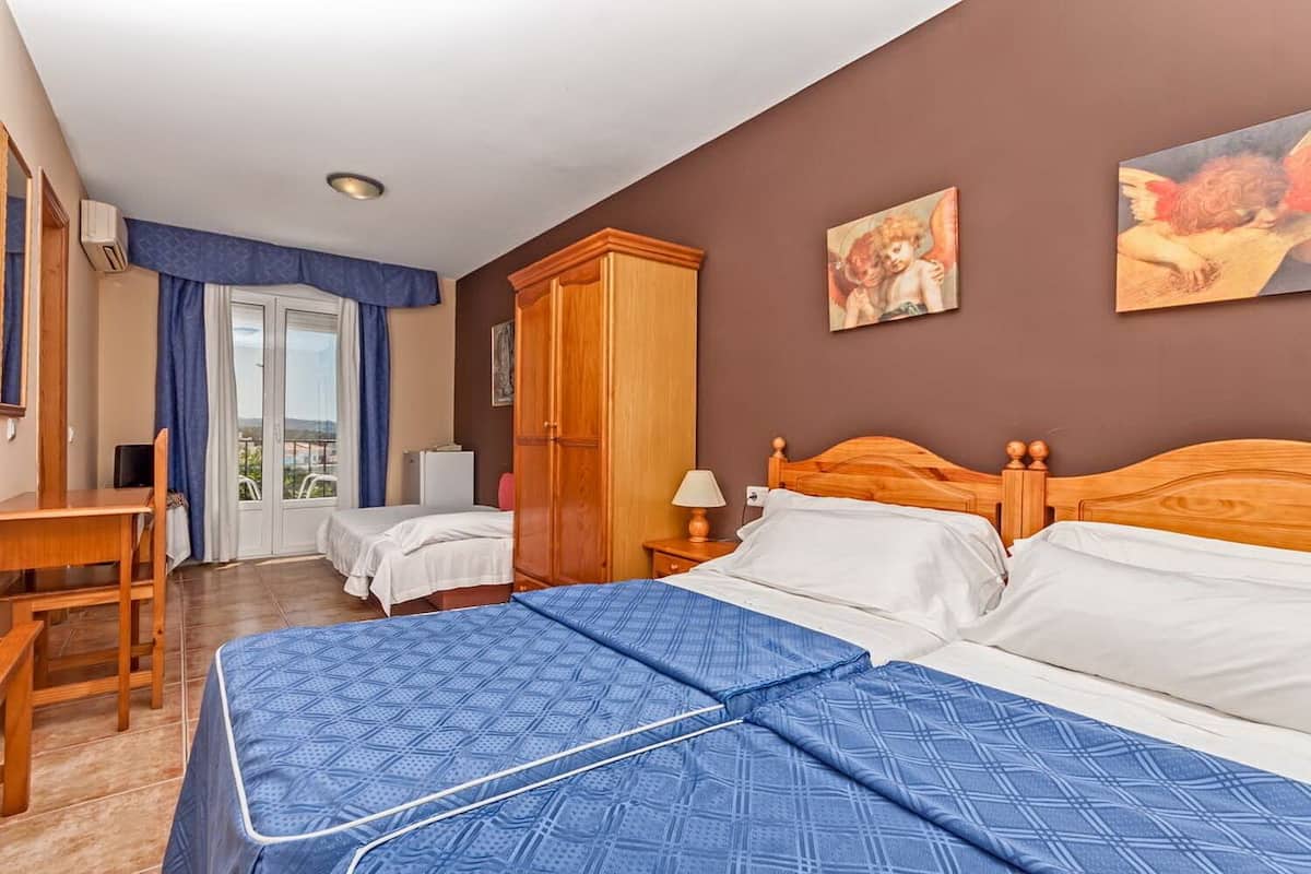 A hotel room with two beds, a desk, a wardrobe, and a view of the outdoors.