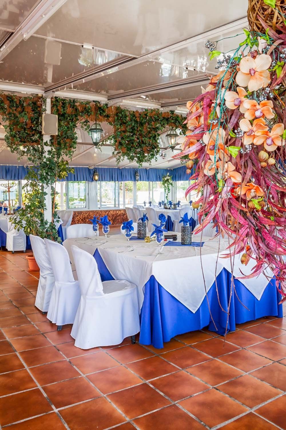 Ein Innenraum mit gedeckten Tischen für eine festliche Veranstaltung, mit weißen und blauen Tischdecken, Stühlen mit weißen Hussen und dekorativen Blumenarrangements.