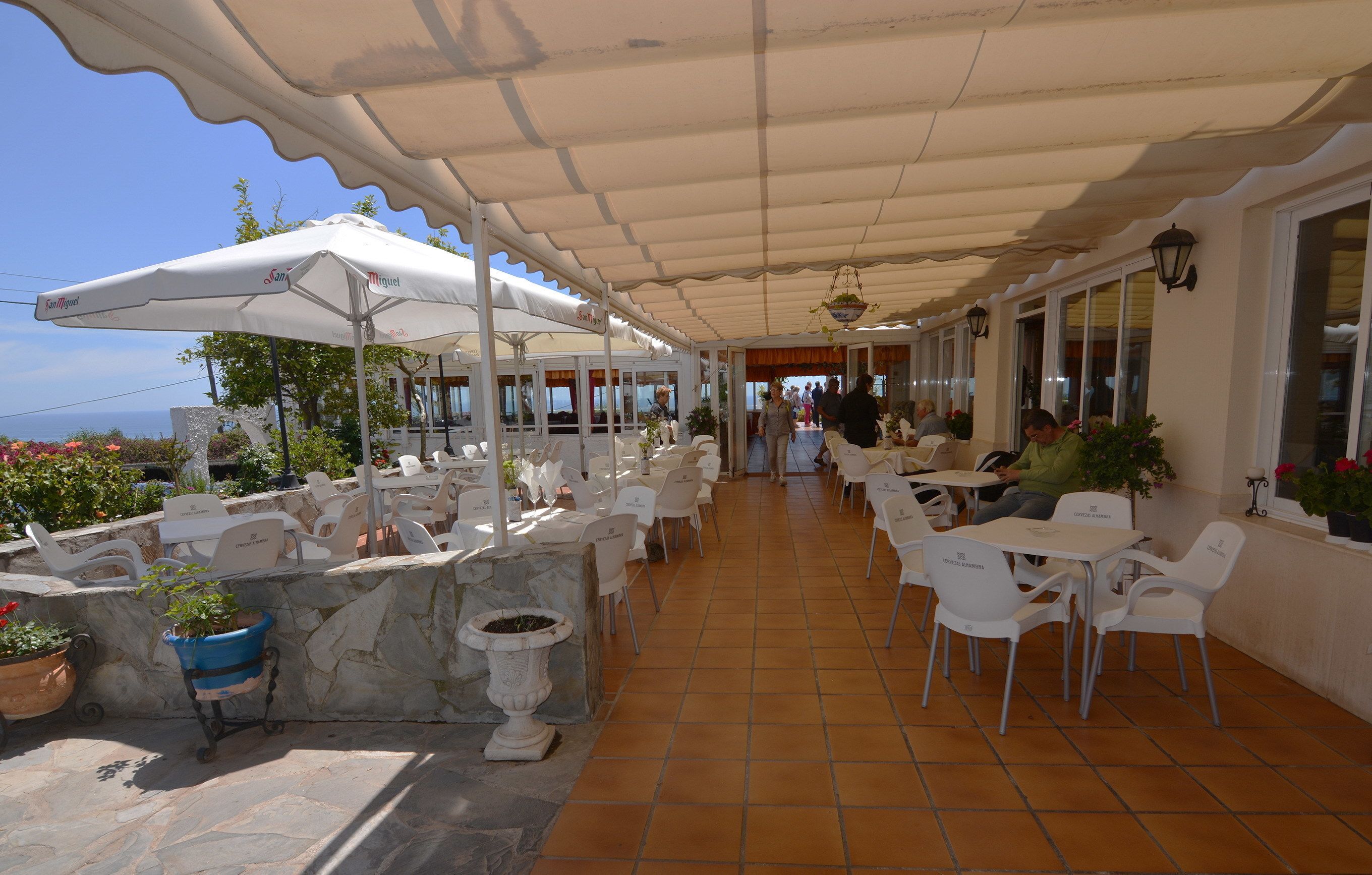 Une terrasse extérieure avec des tables et des chaises blanches, recouverte d’un auvent blanc, et agrémentée de plantes en pot.