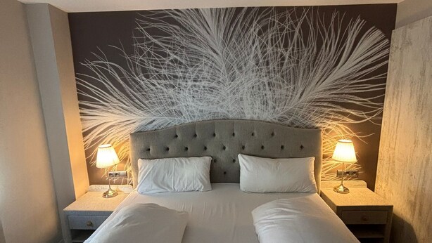 A double bed with a tufted headboard, two bedside tables with lamps, and a decorative wall piece. - Hotel Höhn (Ruedesheim am Rhein)
