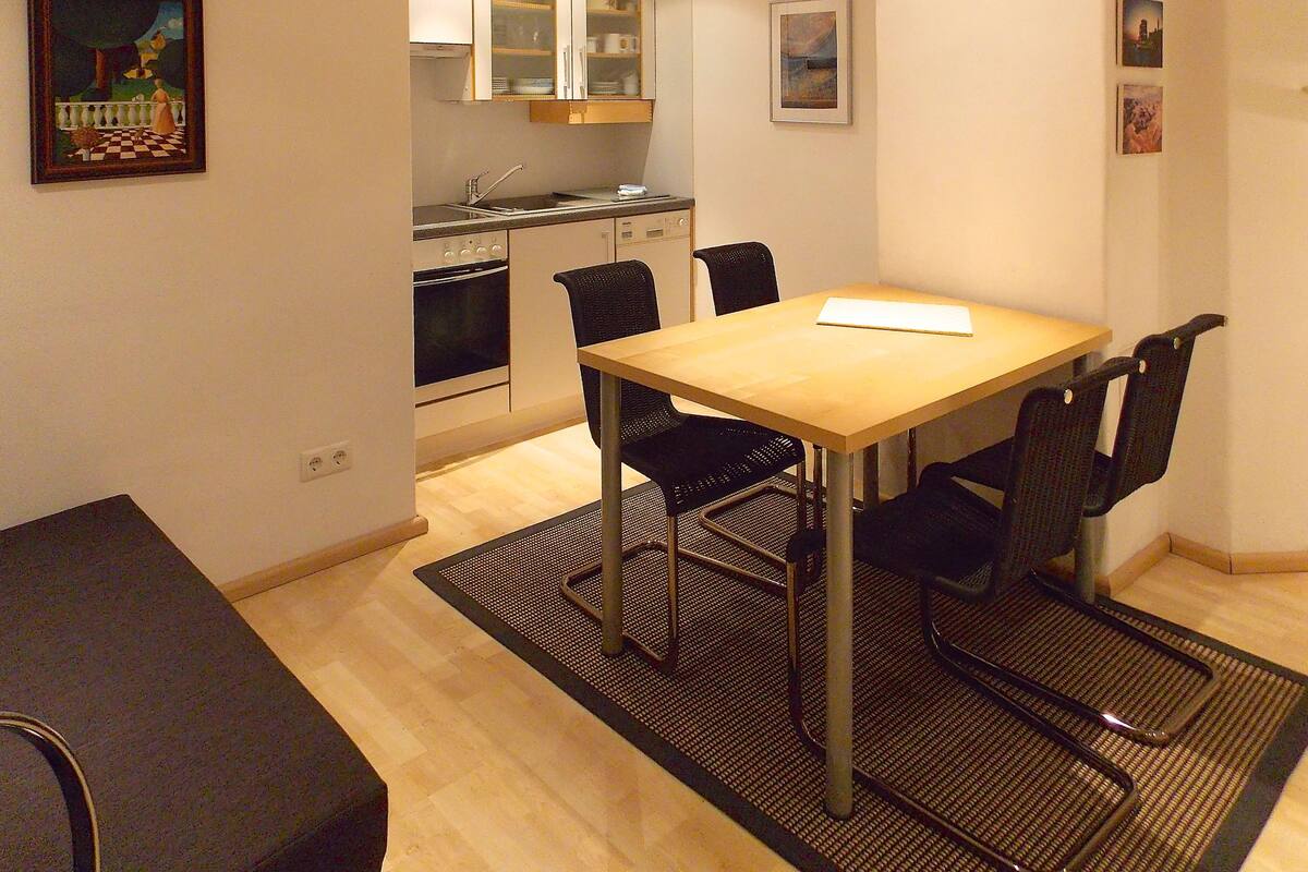 A compact kitchen with a dining area, featuring a wooden table and black chairs, a small oven, and a sink.