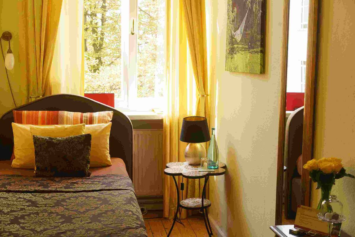 Une chambre d’hôtel avec un lit, une petite table avec une lampe, un vase avec des fleurs et une fenêtre avec des rideaux.