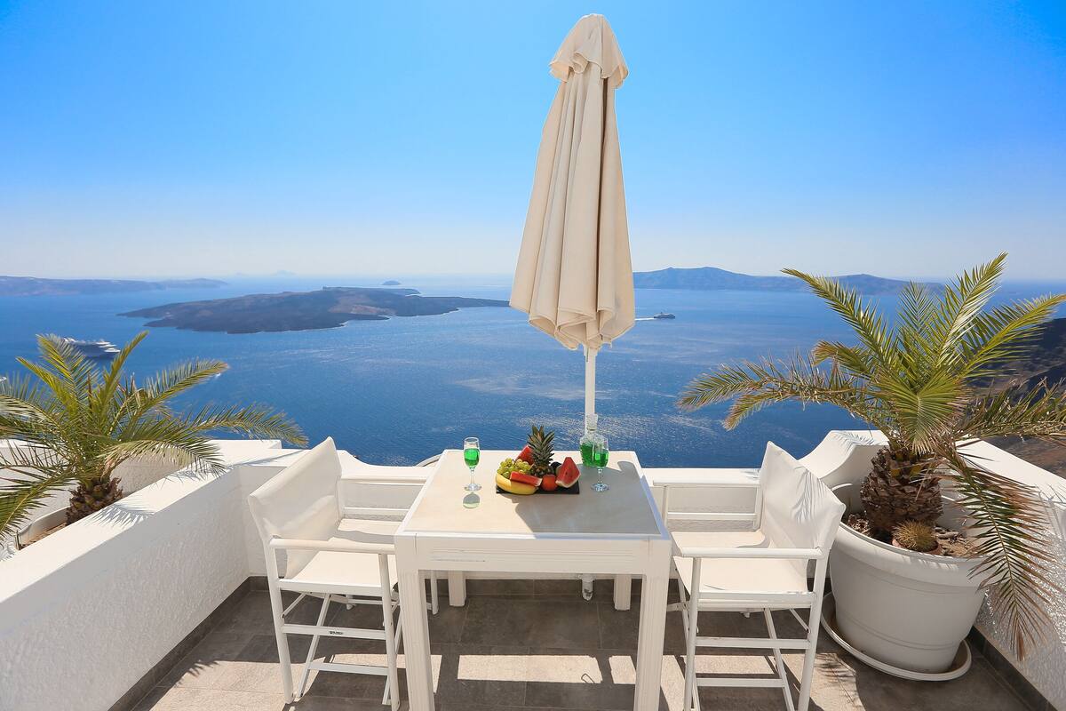 A white table with two chairs and an umbrella, overlooking a scenic view of the sea and islands.