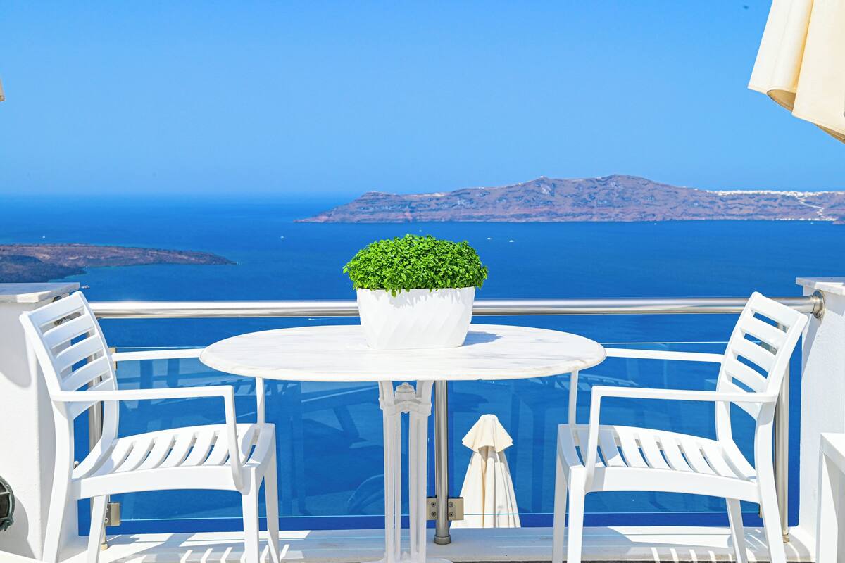 A balcony with a round table and two white chairs, overlooking a clear blue sea and distant land.