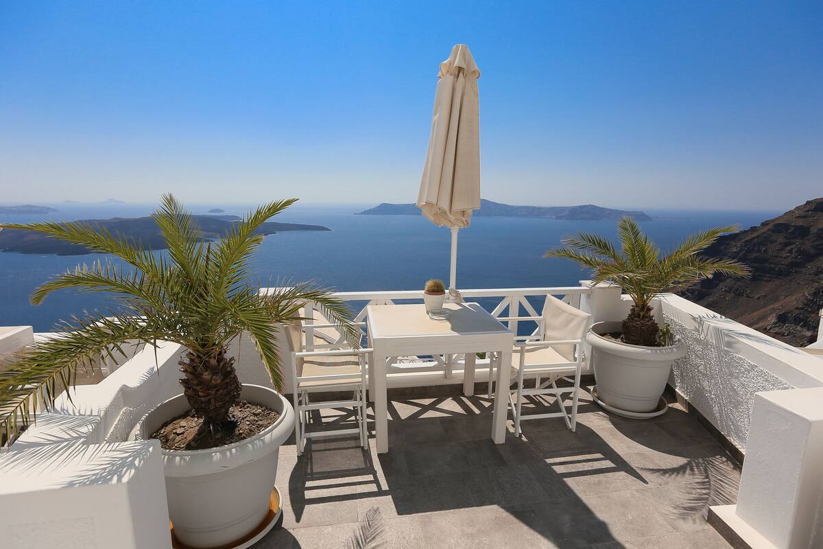 A terrace with a white table and chairs, a potted palm tree, and a view of the sea and distant islands.