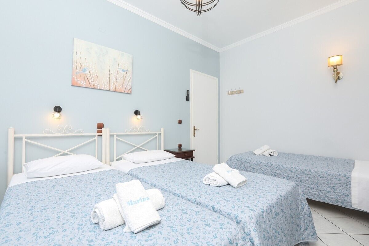 Une chambre d’hôtel avec deux lits, chacun recouvert d’une couvre-lit à motifs floraux bleus et agrémenté d’oreillers blancs. On y trouve des serviettes sur lesquelles est brodé le mot « Marina ».