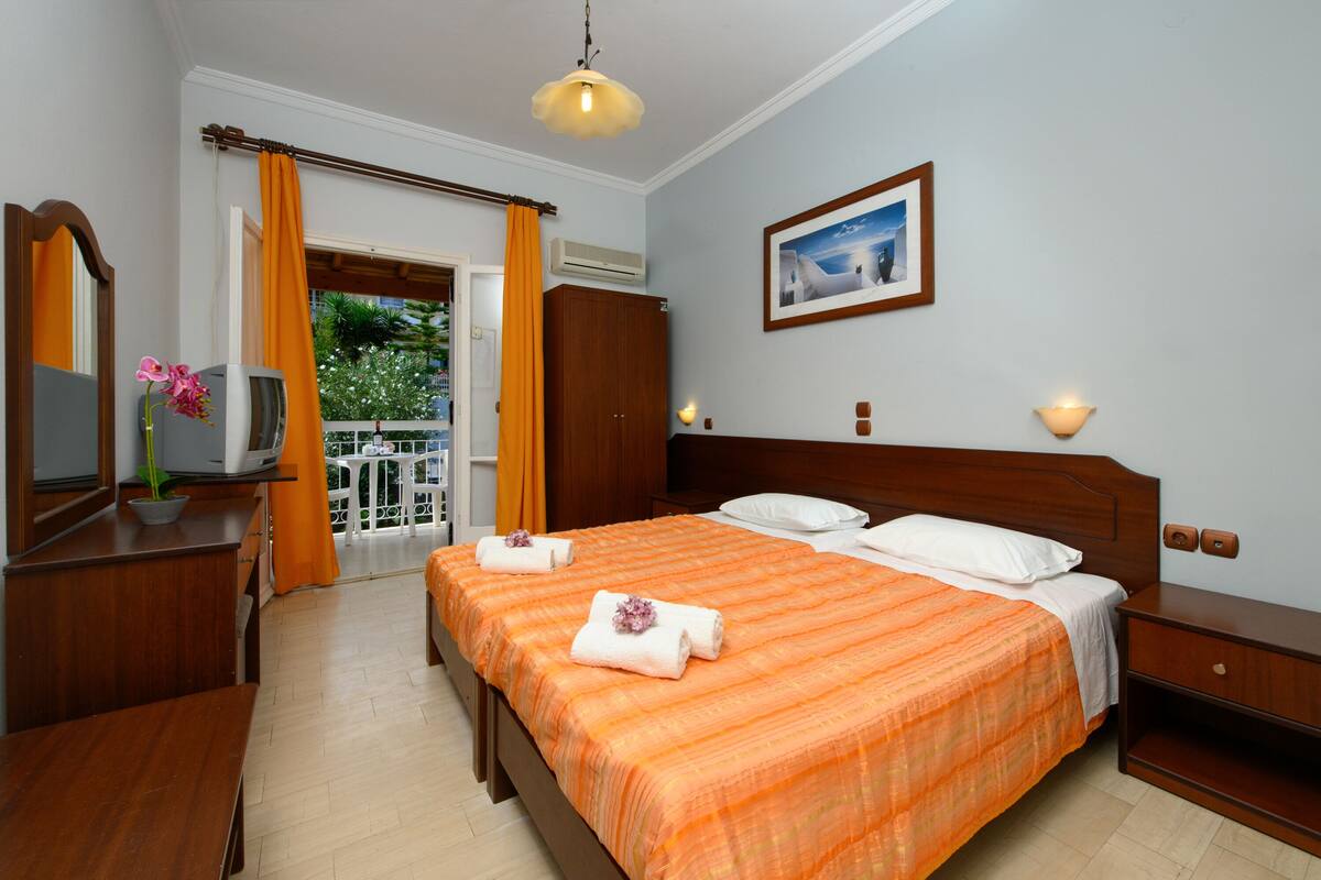 A hotel room with a large bed, a bedside table, a dresser, a mirror, and a balcony with orange curtains.
