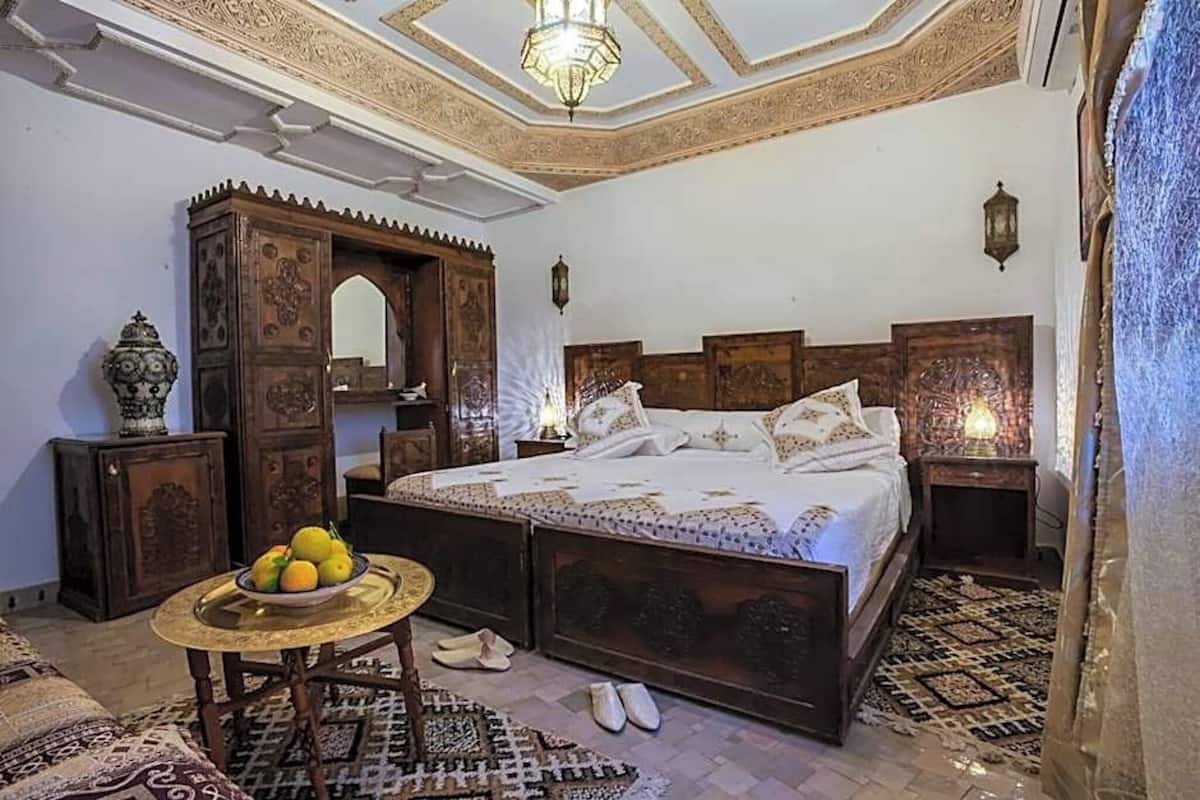 Quarto tradicional com uma cama, uma mesinha com frutas, um espelho e um tapete estampado.