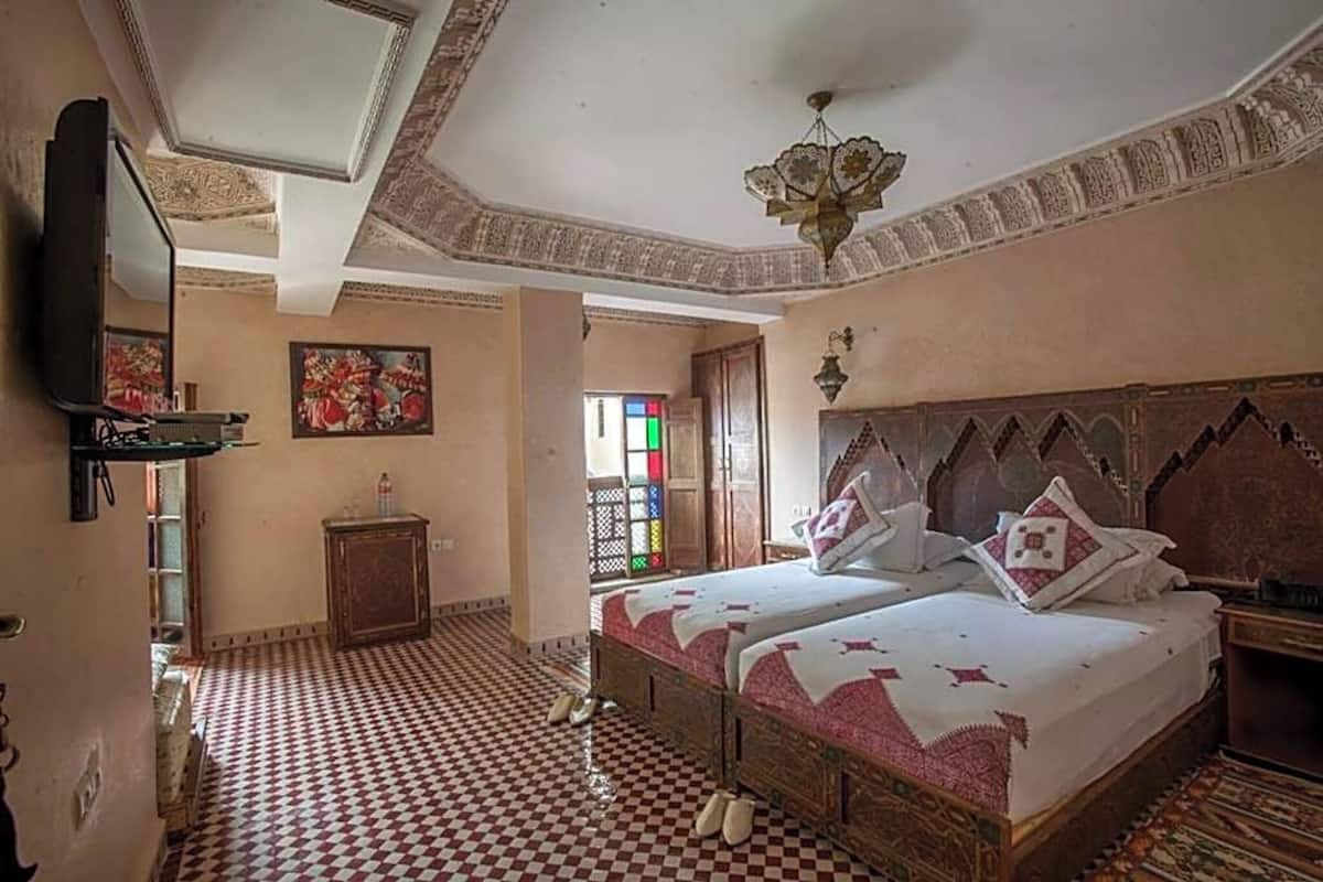 Um quarto com piso quadriculado, uma cama com colcha estampada, um lustre e uma televisão fixada na parede.