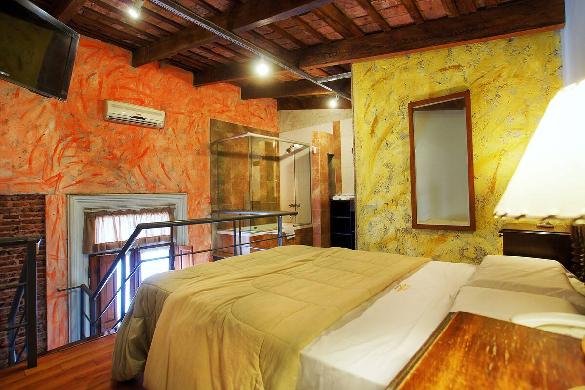 A hotel room with a bed, a wooden floor, a window with curtains, a mirror, and a wall with a yellow pattern.