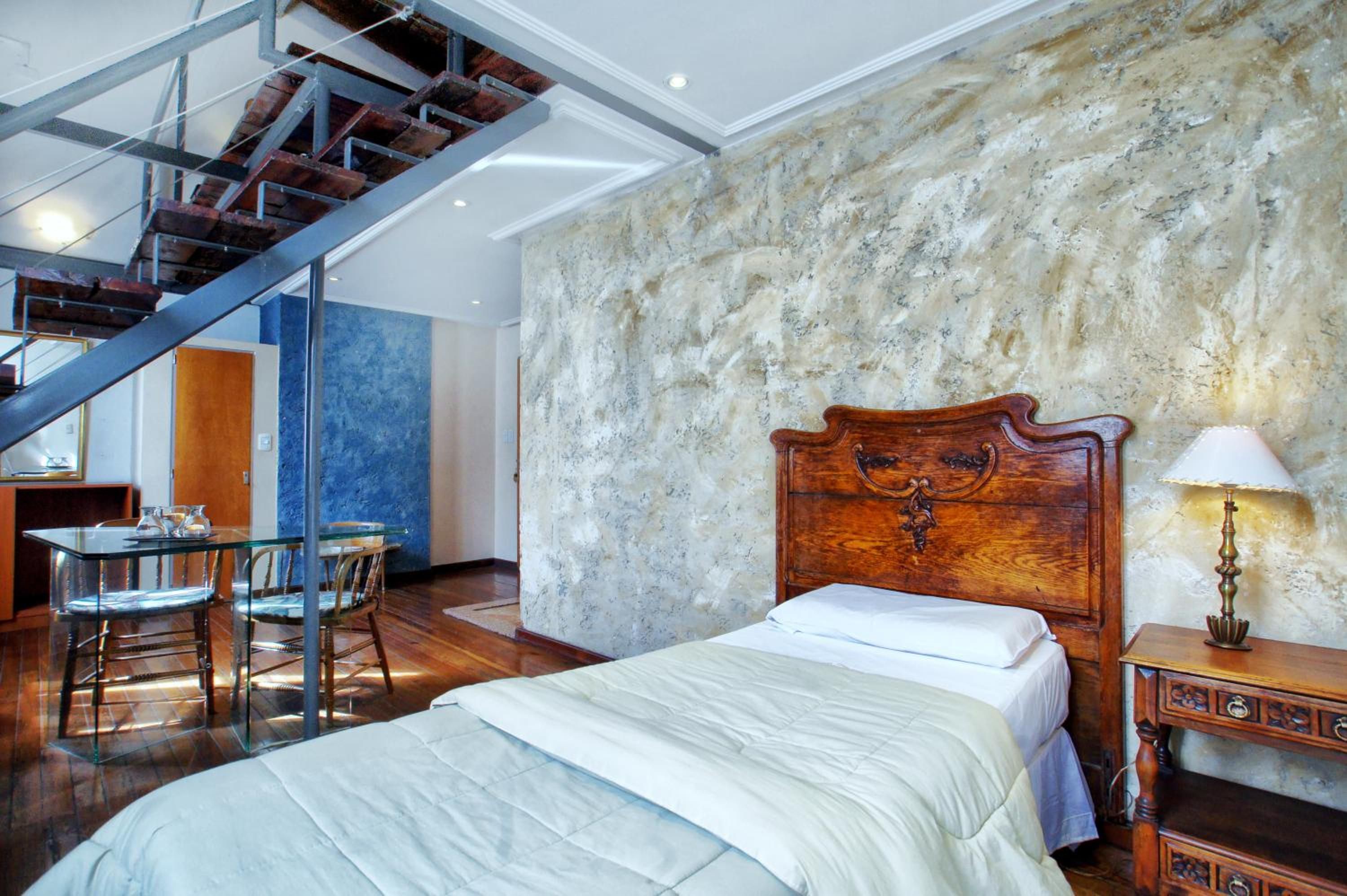 Un dormitorio con cama, mesita de noche, lámpara, silla, mesa y una escalera.
