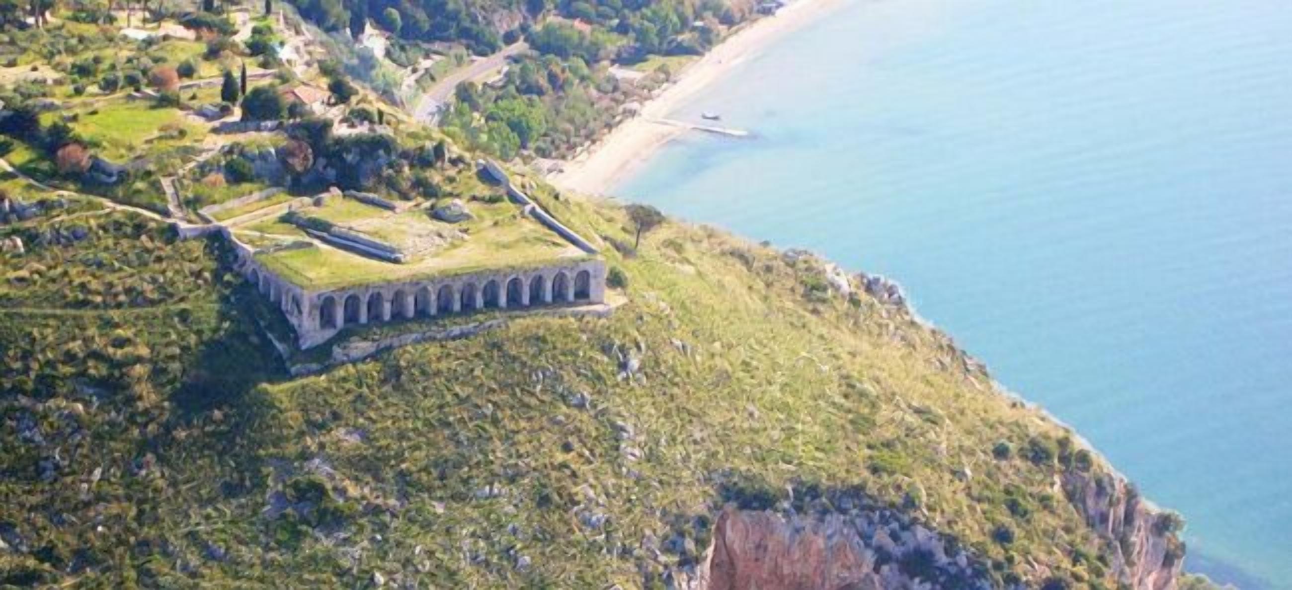 Vista aérea de uma área costeira com um edifício notável, apresentando uma estrutura longa e linear com várias arcos, localizado numa colina gramada com vista para uma praia.