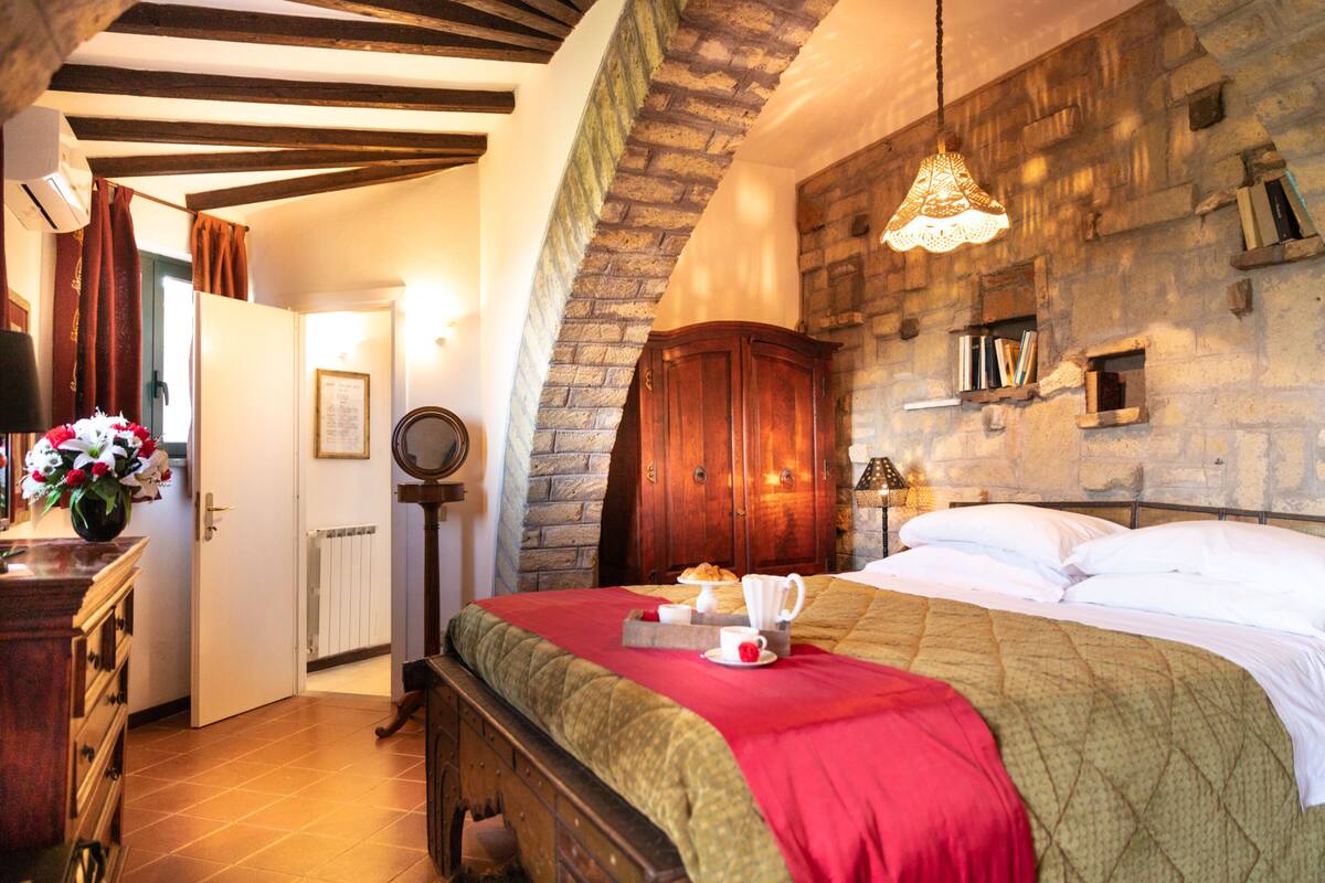 A bedroom with a stone wall, wooden bed frame, and a red bedspread.