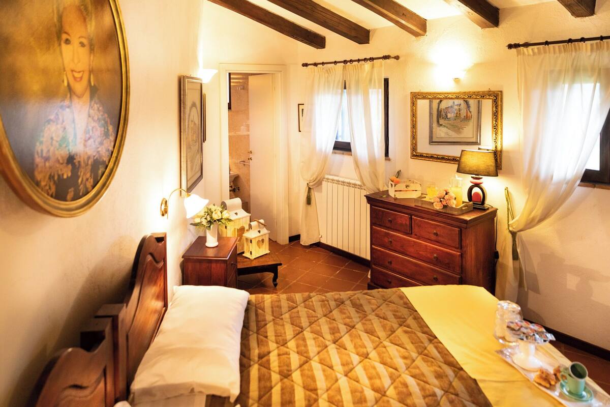 A hotel room with a bed, a wooden dresser, a nightstand, a lamp, a painting, and a window with curtains.
