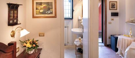 A hotel room with a bed, a desk with a lamp, a framed painting, and a view into the bathroom.