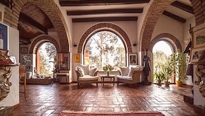 Una habitación amplia con un tapete rojo estampado, dos sillones, una mesa de centro y una estatua. La habitación cuenta con arcos de ladrillo visto y un techo de madera.