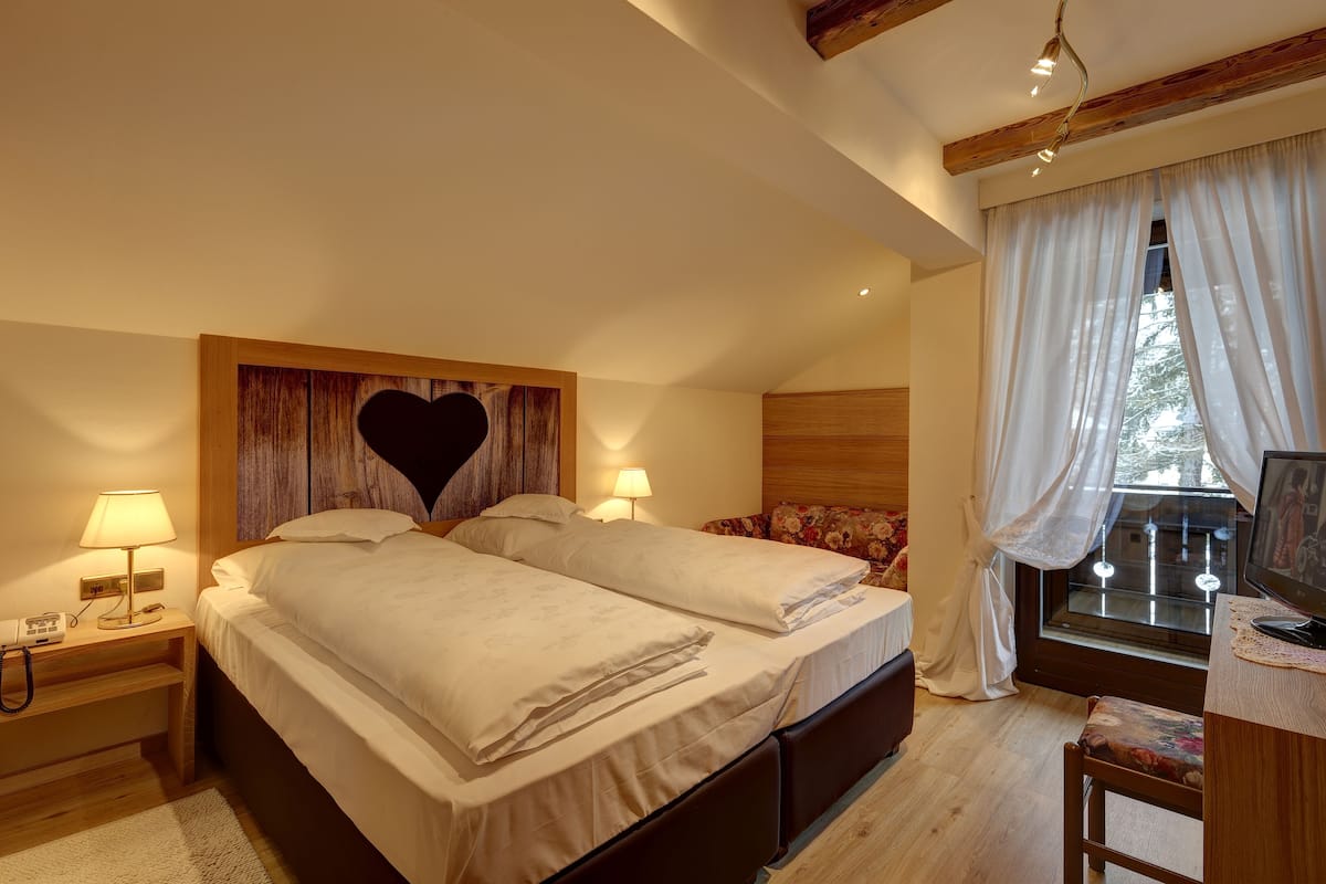 A bedroom with two beds, a wooden headboard with a heart design, bedside lamps, a window with curtains, and a desk with a computer.
