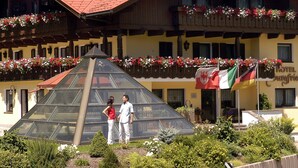 Un hotel de estilo arquitectónico alpino tradicional, con una estructura de vidrio piramidal destacada en primer plano, rodeado de exuberante vegetación y jardineras florales.