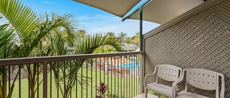A balcony with two white plastic chairs, a tiled floor, a metal railing, and a view of a pool and lawn.