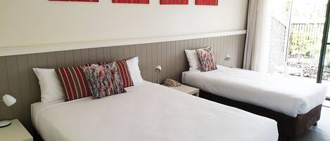 A hotel room with two beds, a small table, a bedside lamp, and a window with a view of the outside.