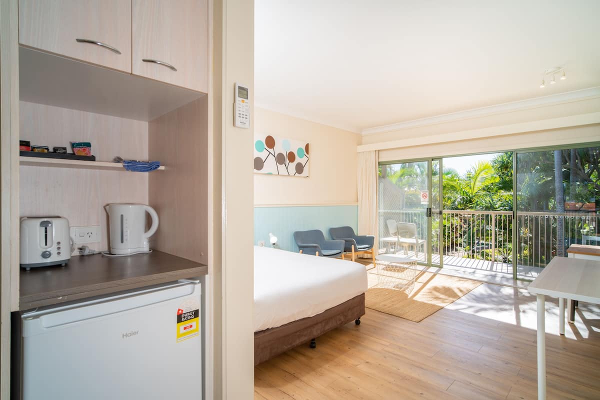 A hotel room with a bed, a kitchenette, a balcony with chairs, and a view of greenery.