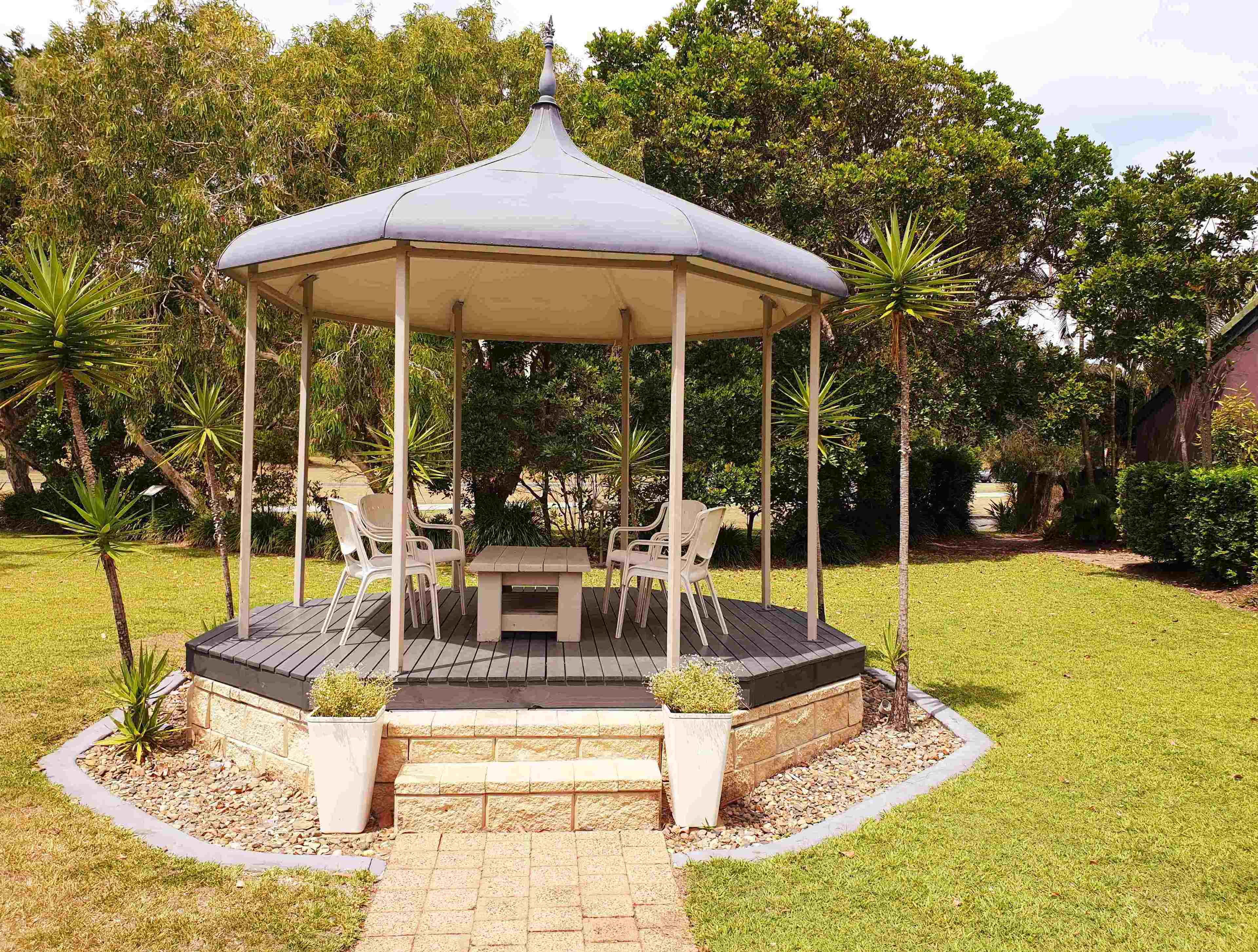 A gazebo with a table and chairs, surrounded by a garden with various plants and trees.