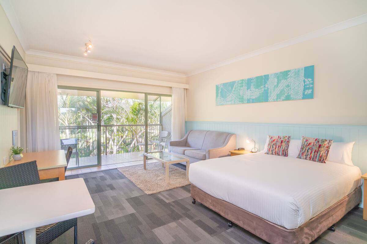 A hotel room with a large bed, a dining table, a sofa, and a balcony with a view of palm trees.