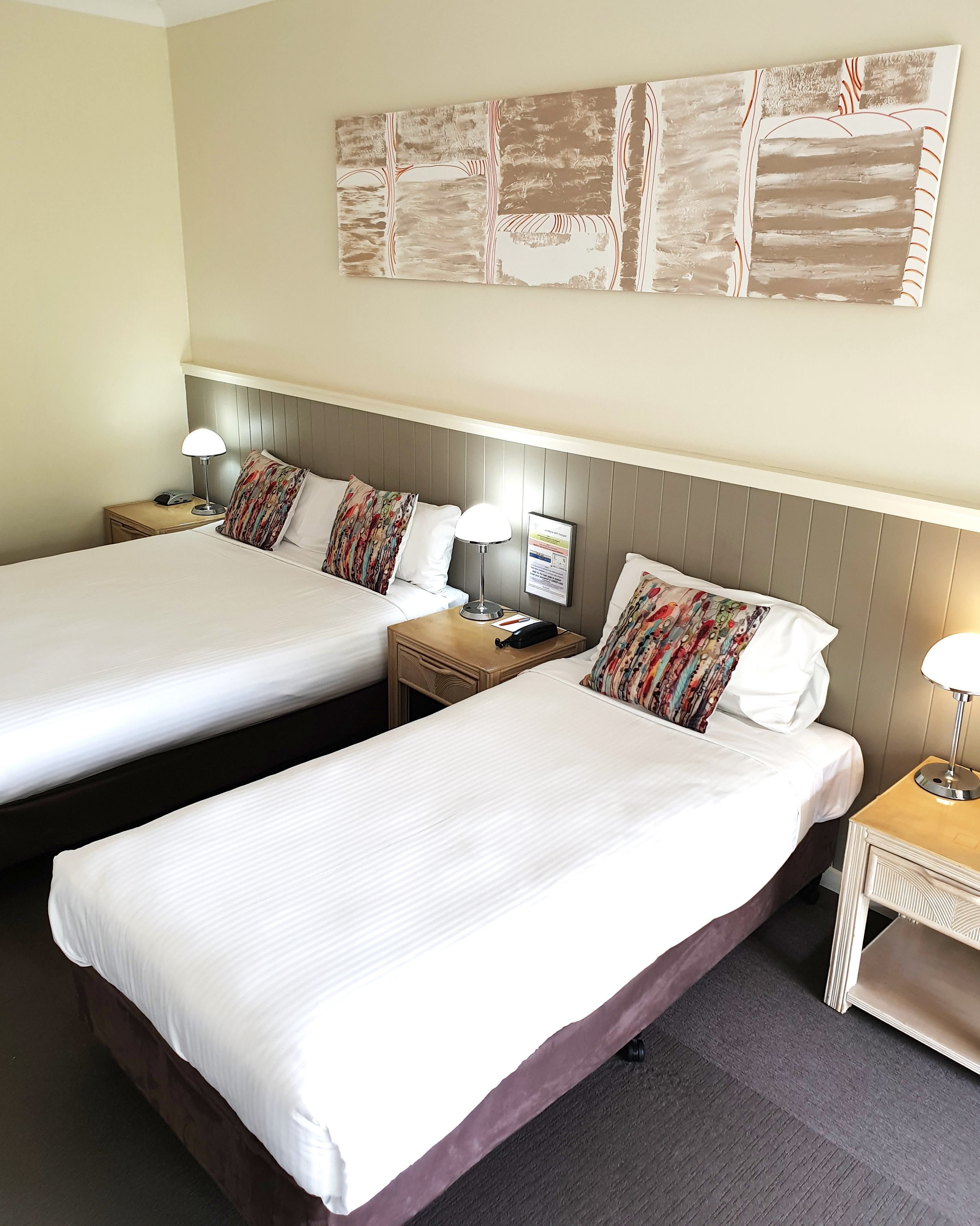 Ein Hotelzimmer mit zwei Einzelbetten, jeweils mit Nachttisch, Lampe und Telefon ausgestattet. Das Zimmer hat eine neutrale Farbgebung mit beigen Wänden und einem großen, abstrakten Kunstwerk über den Betten.
