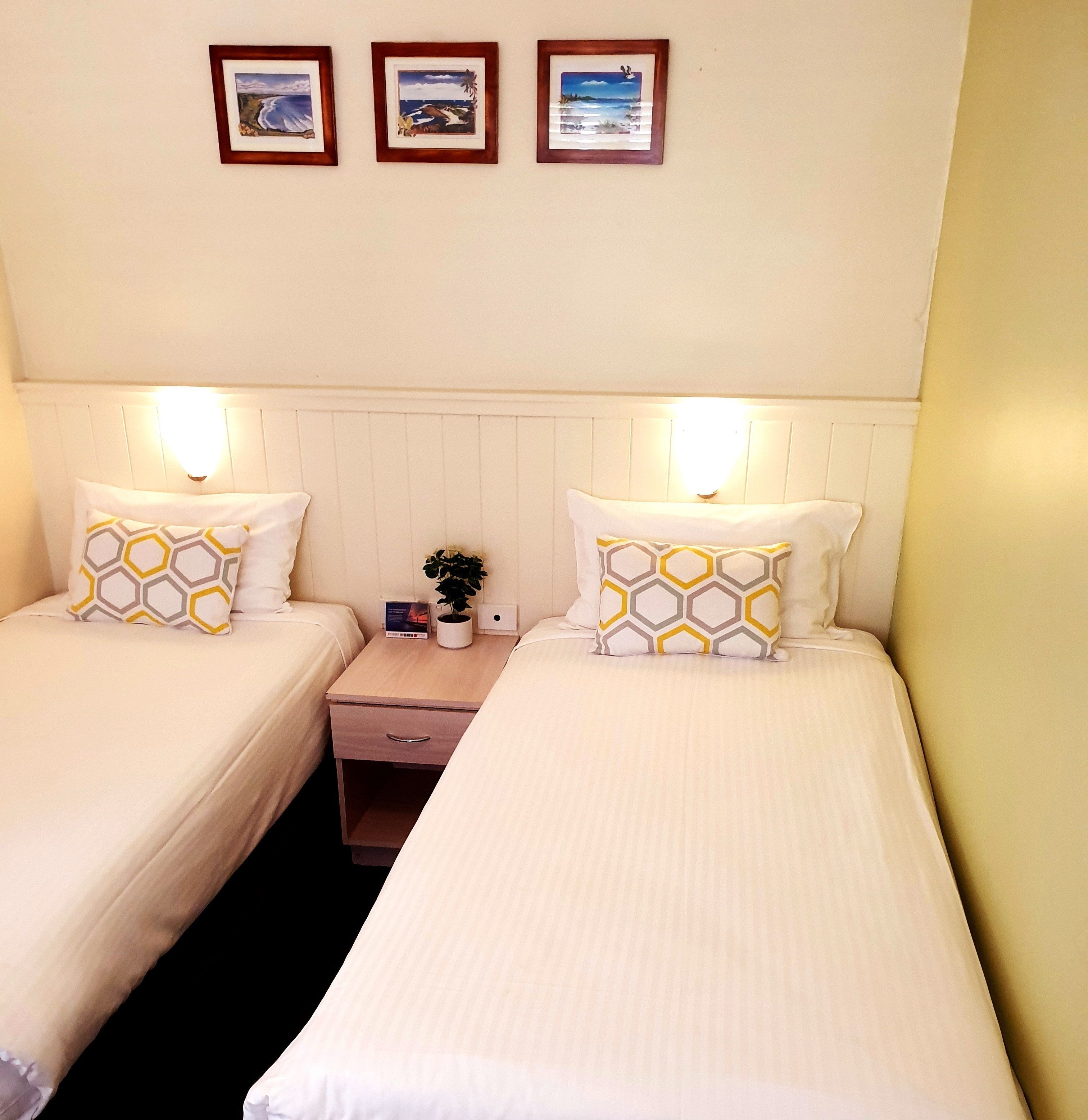 Een hotelkamer met twee bedden, elk voorzien van een witte spreidek en passende kussens. Boven de bedden hangen drie ingekaderde foto's.