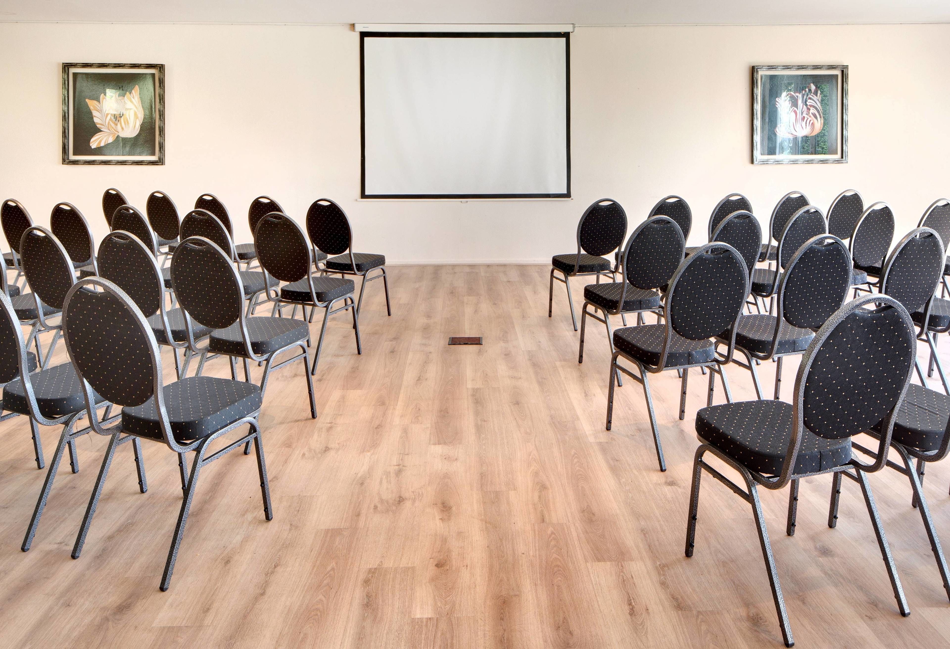 A conference room with rows of chairs facing a blank screen, wooden flooring, and two framed artworks on the walls.