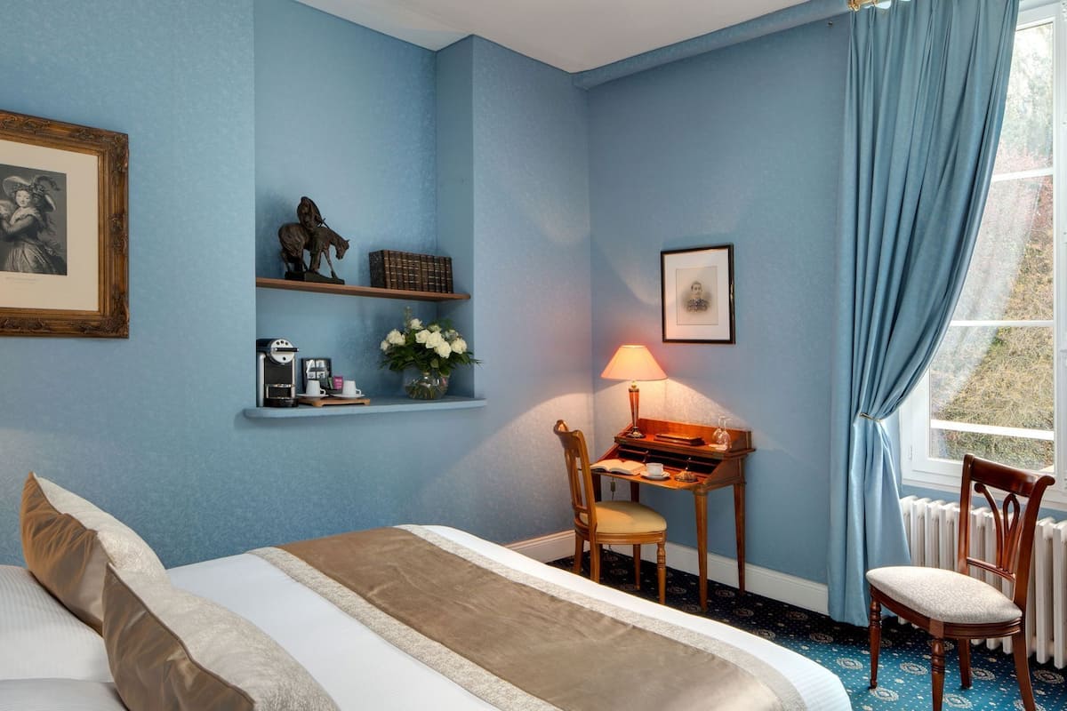 Un dormitorio con una cama, un escritorio con lámpara, una silla, una estantería con objetos decorativos y una ventana con cortinas.