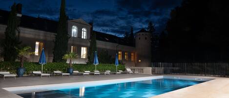 Una piscina con sillones de descanso y sombrillas, rodeada por un edificio con una torre y un seto.