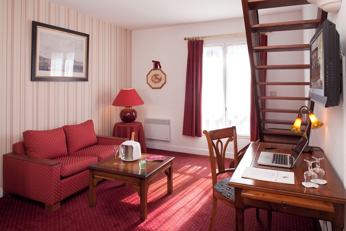 Una habitación acogedora con un sofá rojo, una mesa de madera con una computadora portátil, una mesita con un tostador y una lámpara roja.