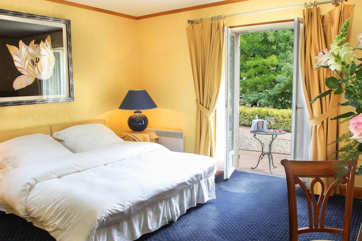 Una habitación de hotel con una cama grande, una mesita de noche con lámpara, una silla y vistas a un jardín a través de una puerta abierta.