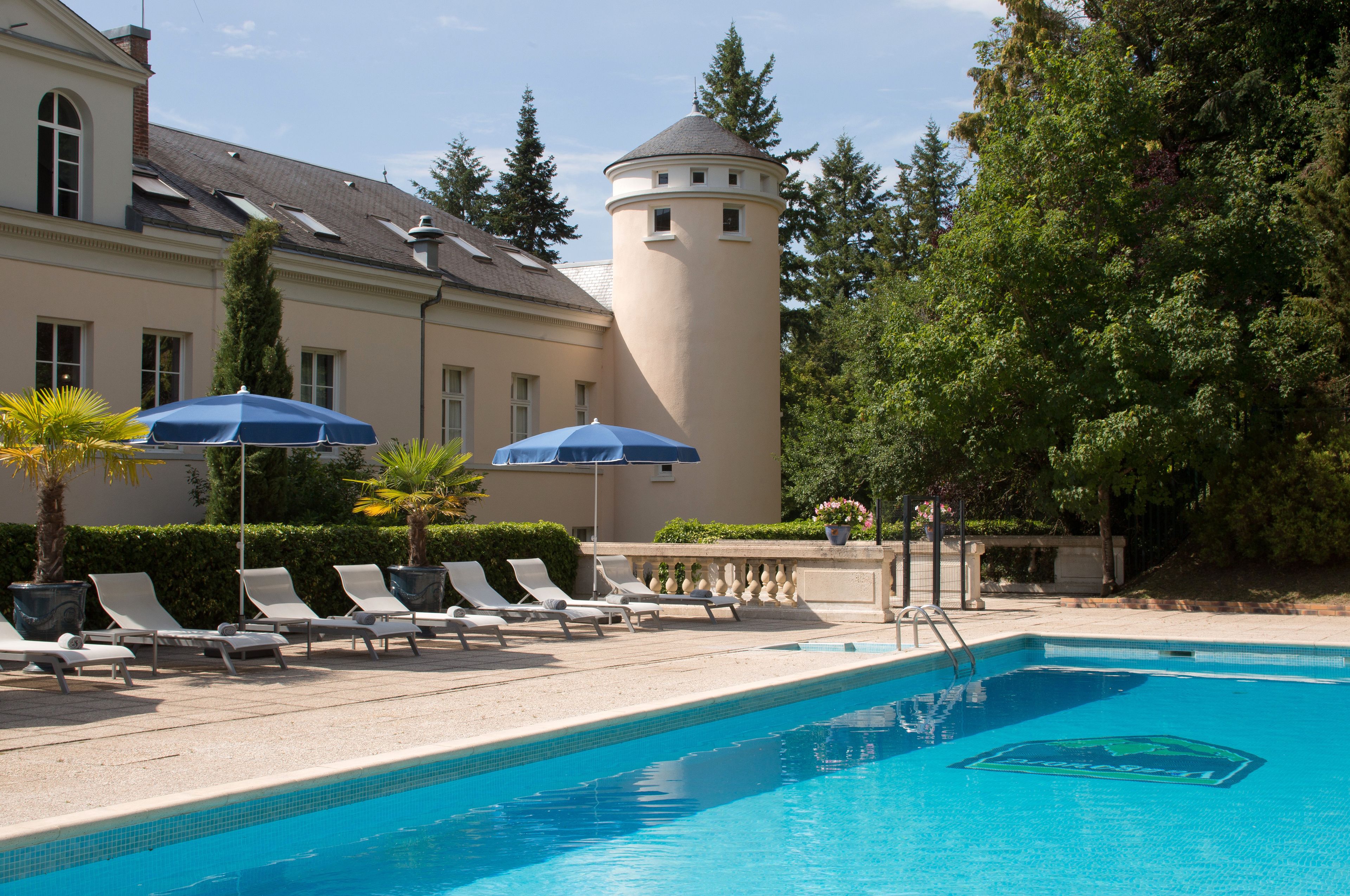 Une piscine avec des chaises longues et des parasols, une tour et un bâtiment surmonté d’une tourelle.