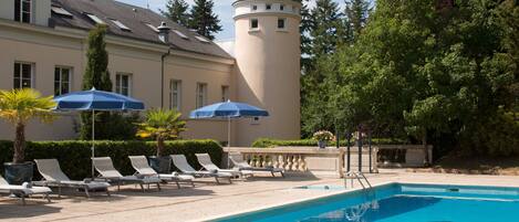 Une piscine avec des chaises longues et des parasols, une tour et un bâtiment surmonté d’une tourelle.