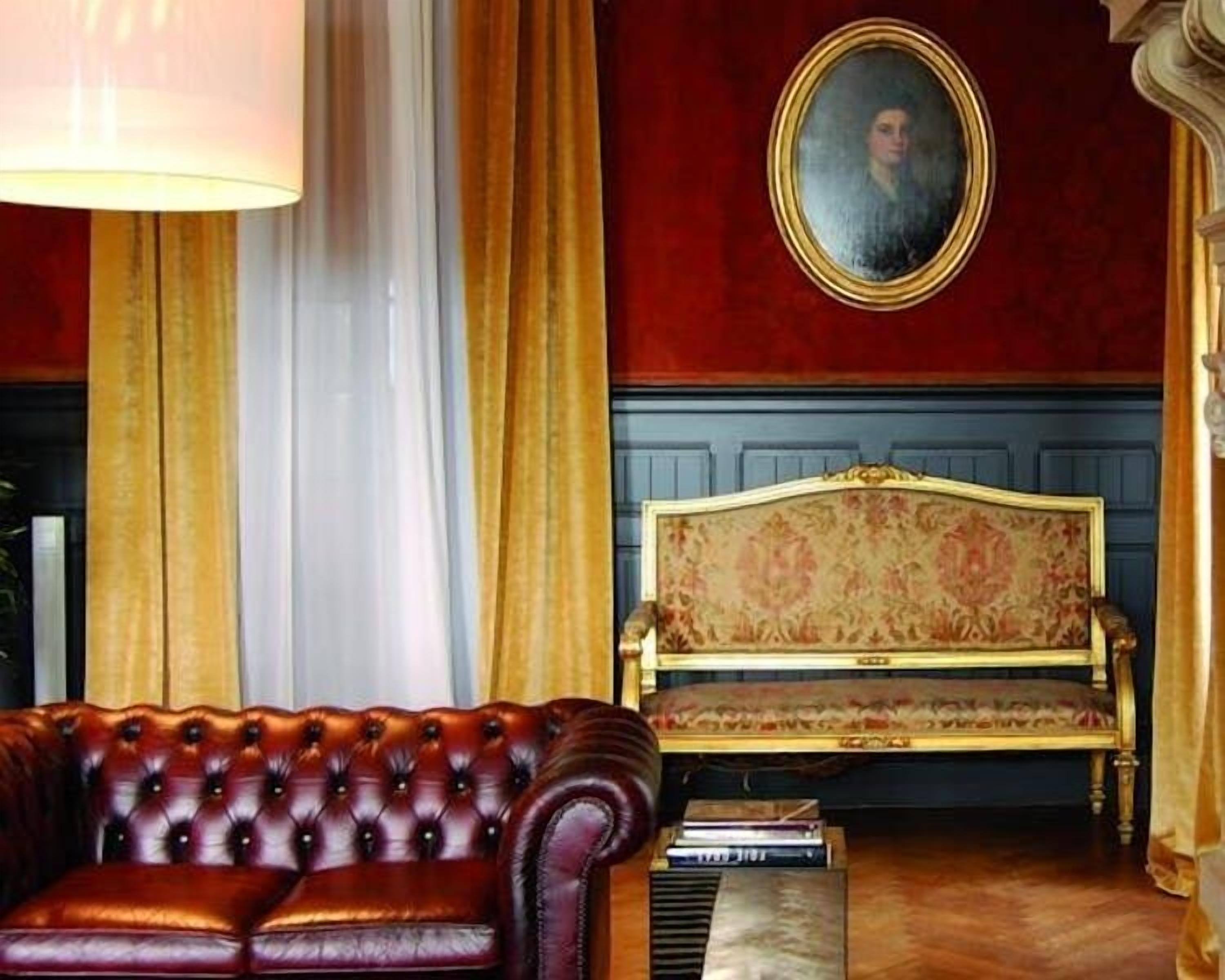 A room with a dark brown leather sofa, a gold-framed sofa with floral upholstery, a portrait on the wall, and a lamp.