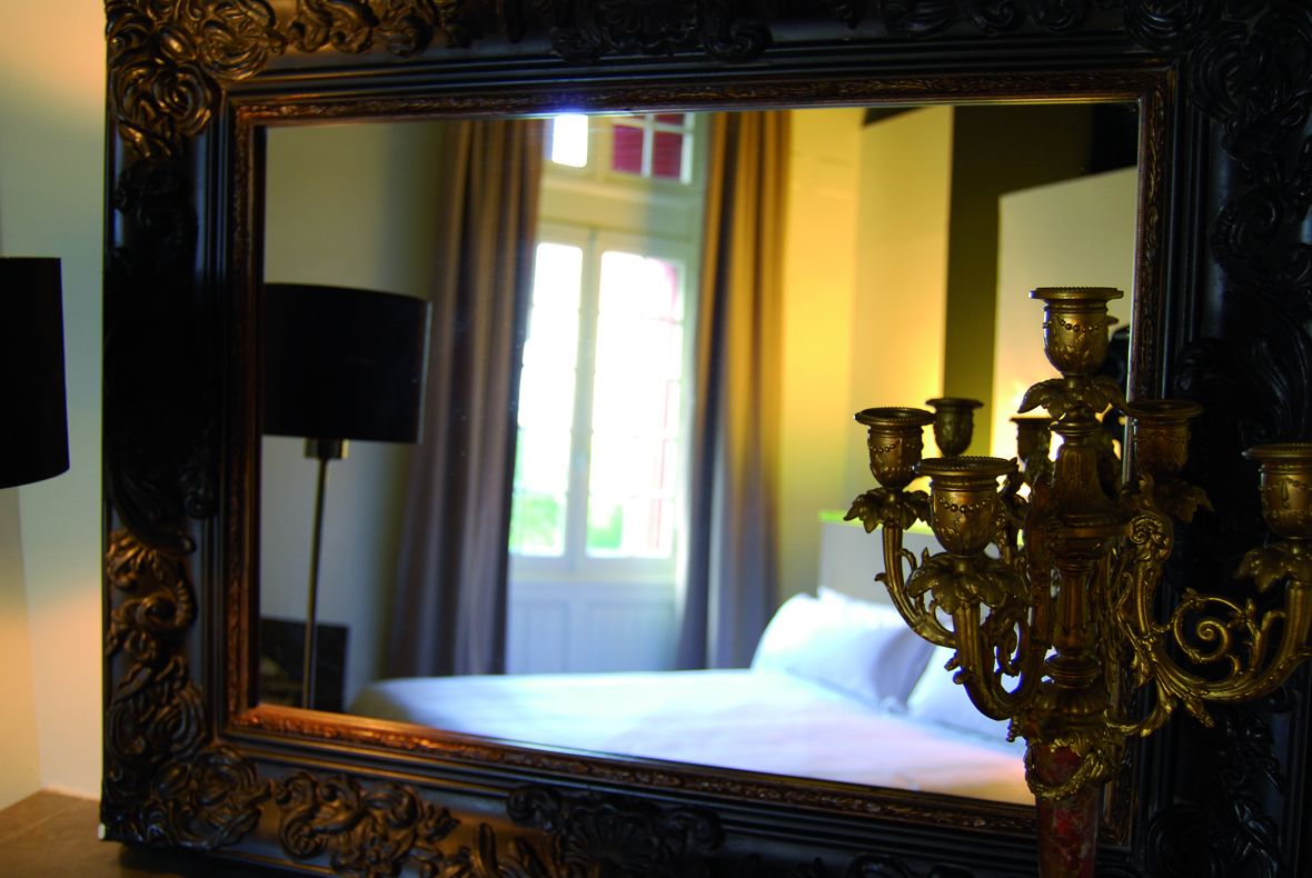 A mirror with an ornate frame reflecting a bed and a window with curtains.