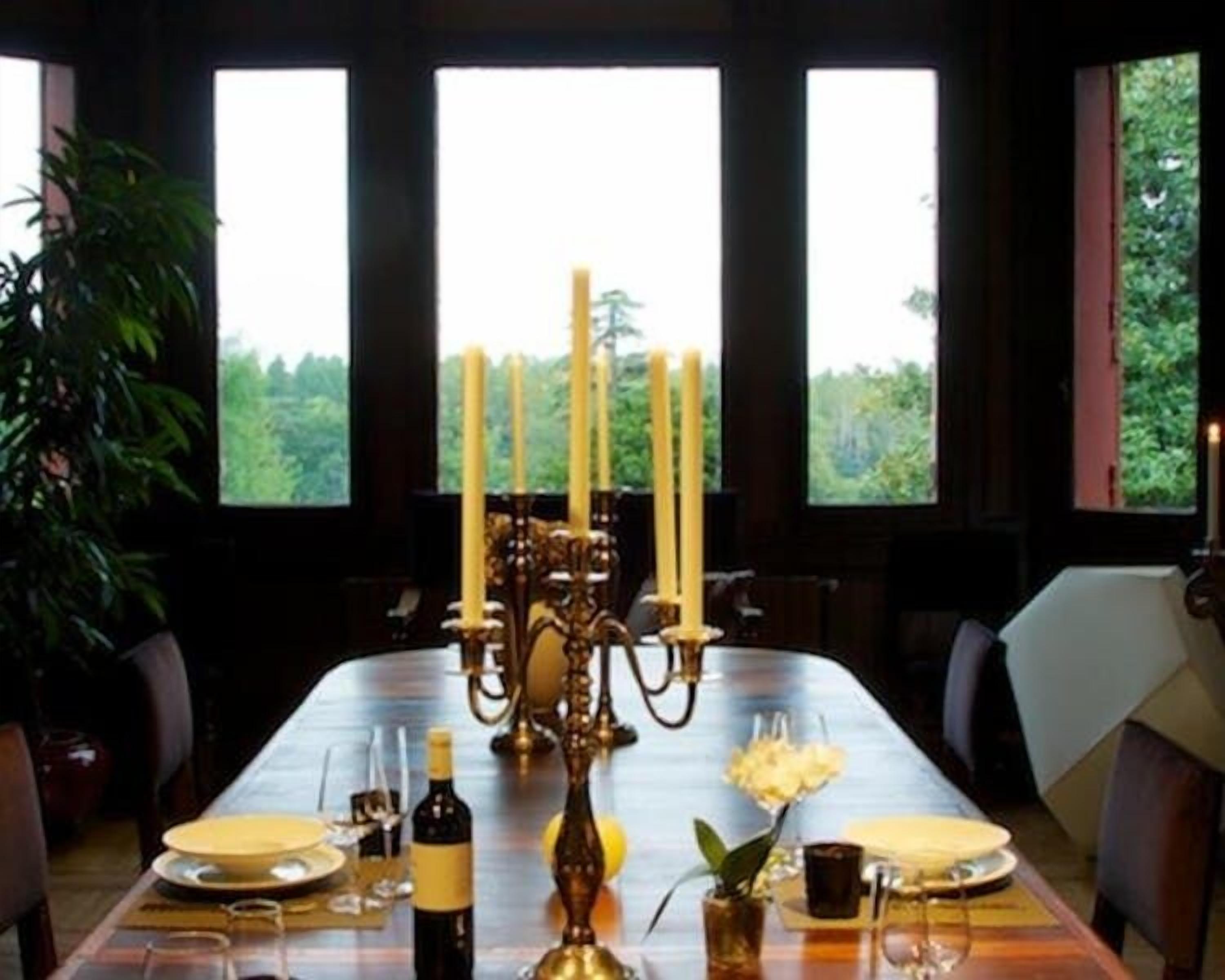 A dining table set with candles, plates, and a bottle of wine, with a view of greenery through large windows.