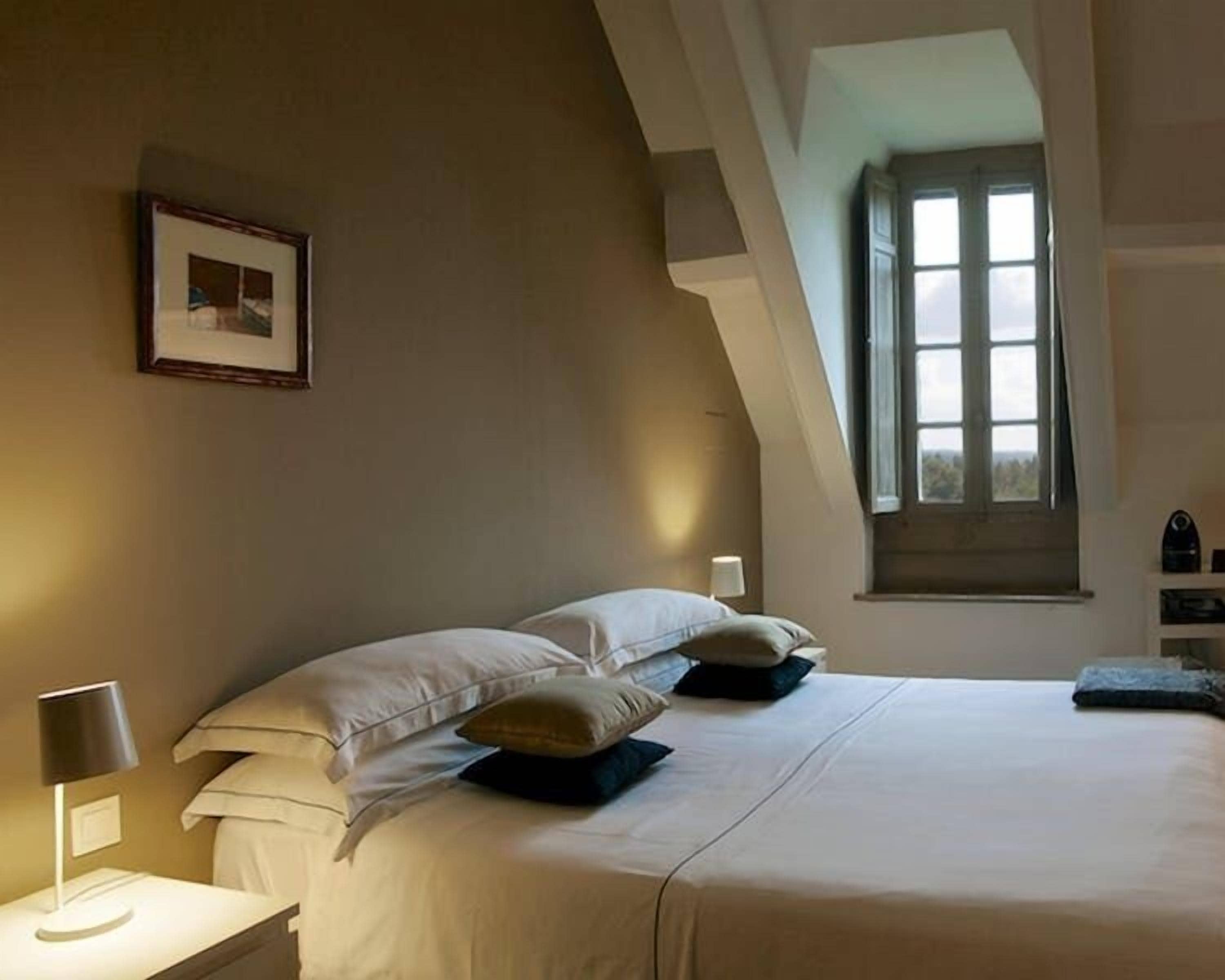Un dormitorio con una cama grande, una mesita de noche con lámpara, un cuadro enmarcado en la pared y una ventana con cortinas.