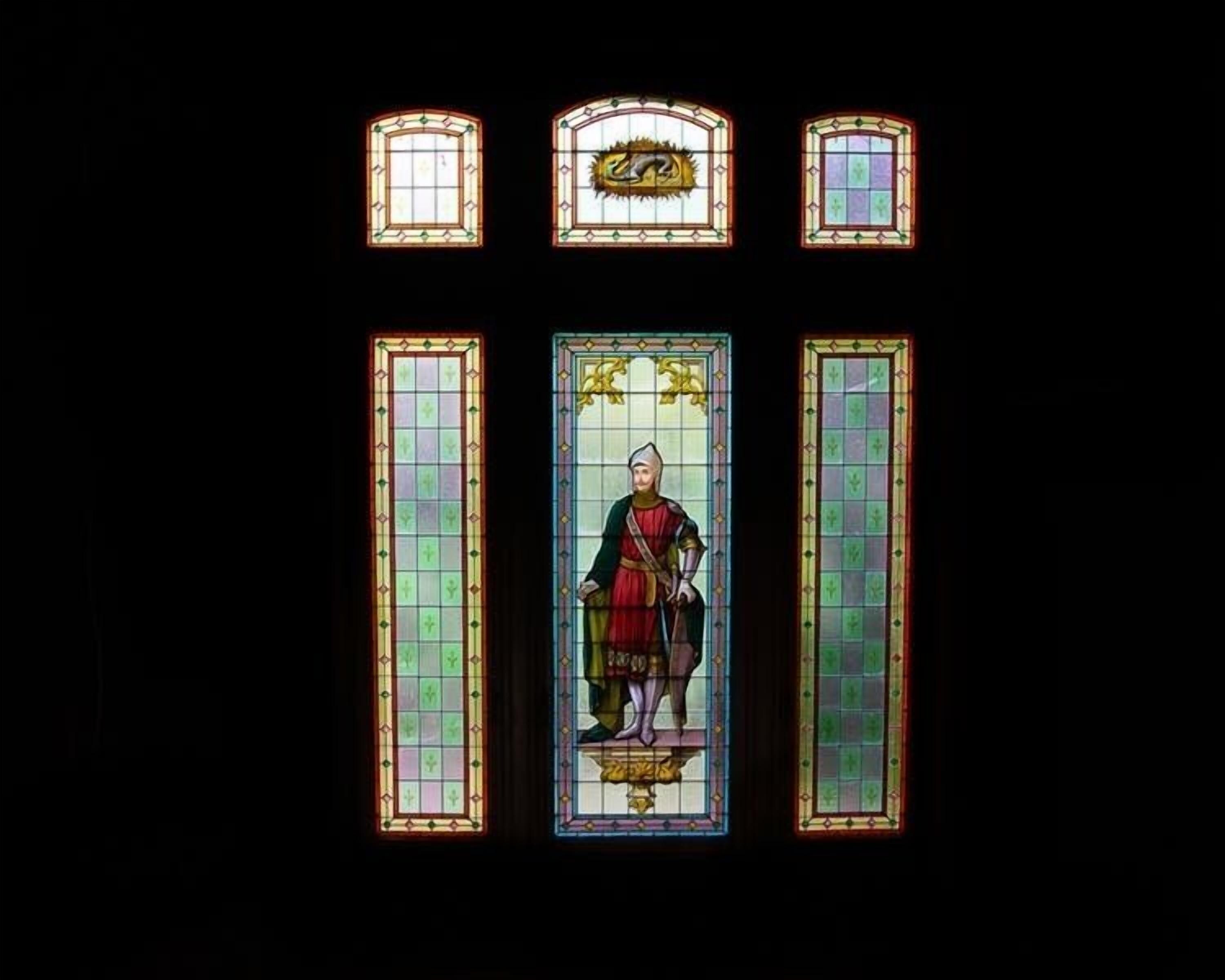 A stained glass window with a central figure in historical attire, flanked by smaller windows with geometric patterns.
