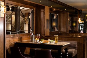 Interior de madera con una mesa dispuesta para comer, incluyendo una botella de vino, una copa de vino tinto y una jarra de cerveza.