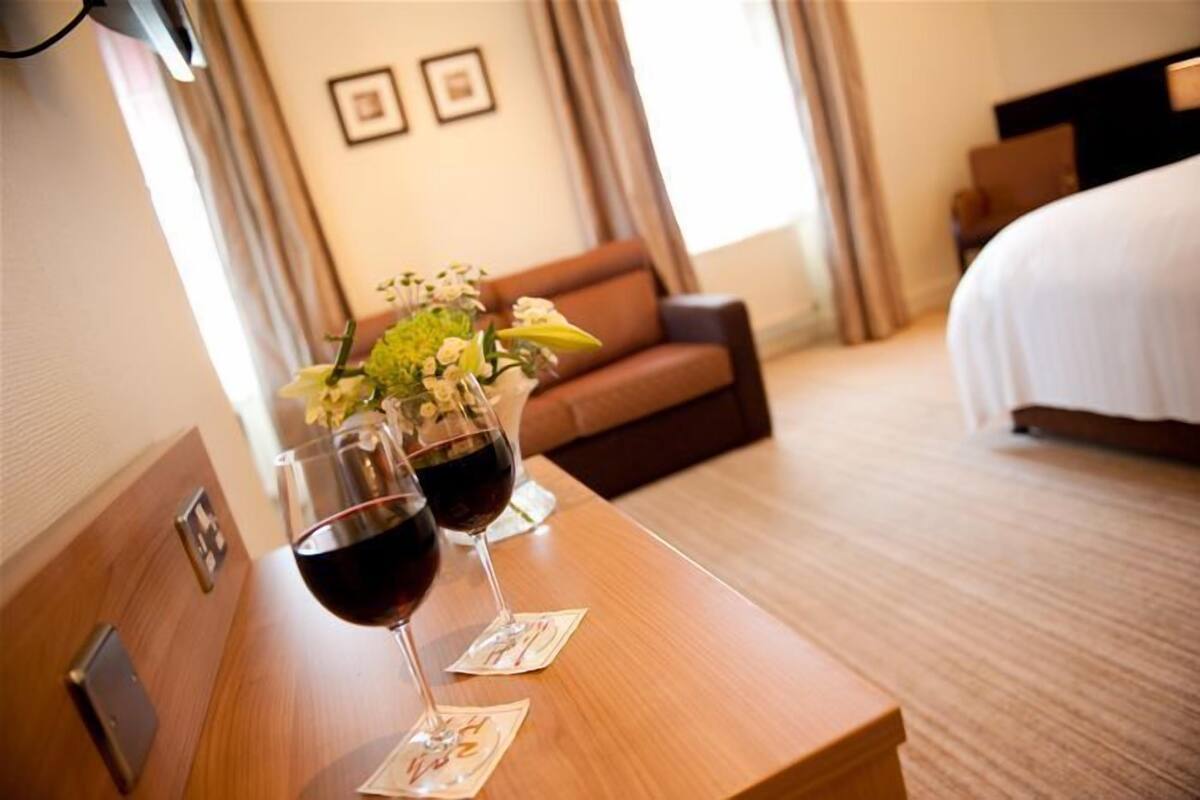 Habitación de hotel con un escritorio de madera, dos copas de vino, un florero con flores, un sofá marrón y una cama con ropa blanca.