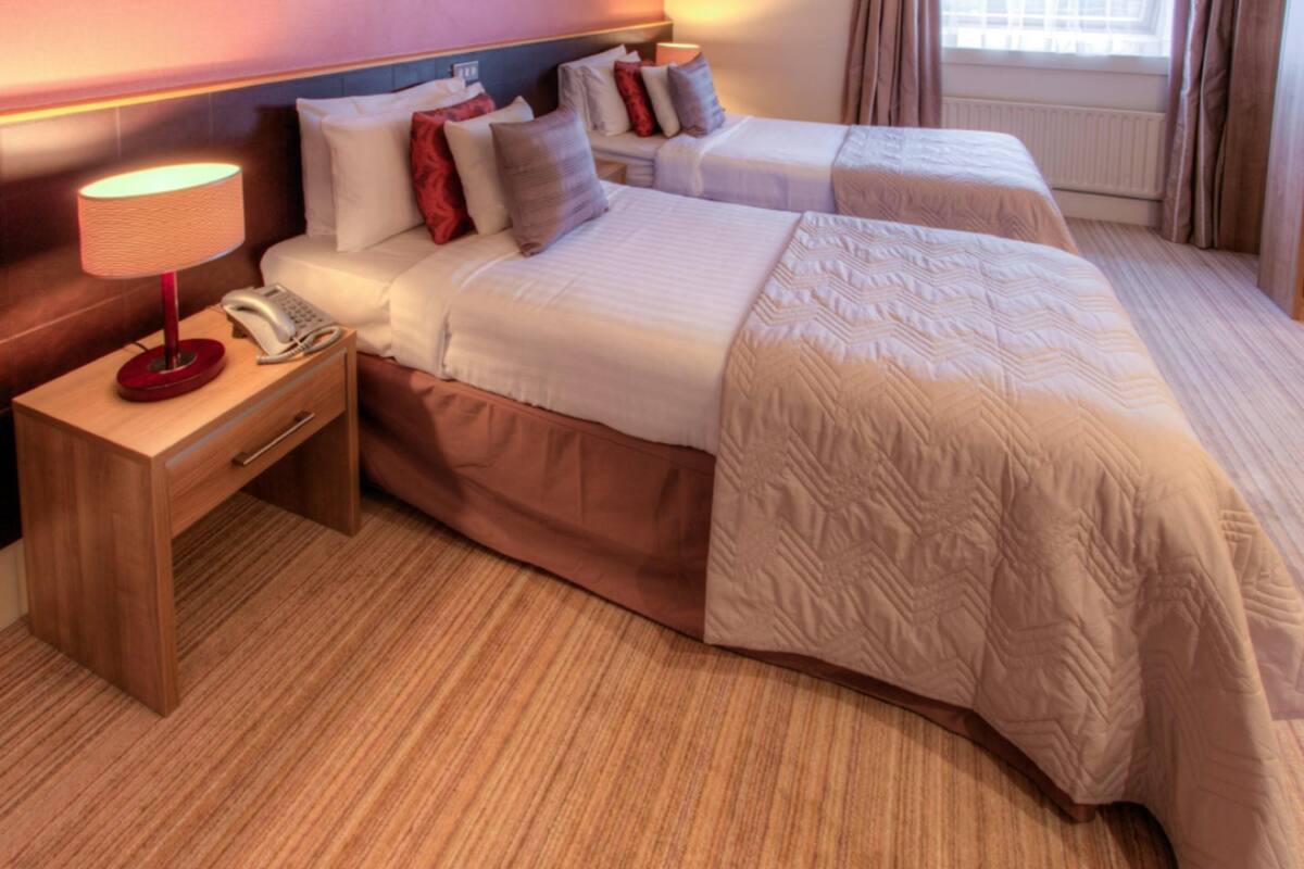 Habitación de hotel con dos camas, una mesita de noche de madera con lámpara y teléfono, y una ventana con cortinas.