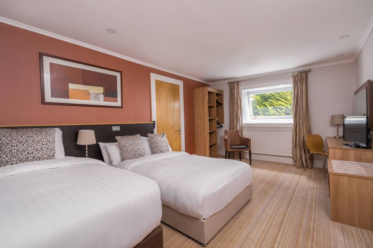 Habitación de hotel con dos camas, un escritorio con televisor, una silla, un armario y una ventana con cortinas.