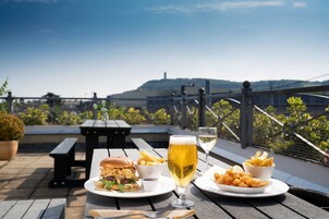 Una mesa de comedor con una hamburguesa, papas fritas y una cerveza en una terraza con vistas a colinas.