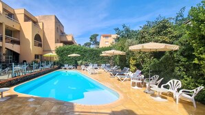Piscina con sillas de descanso y sombrillas, rodeada por un edificio con balcones y un cielo despejado.